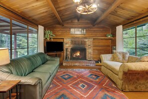 A wooden interior with a fireplace, two sofas, a TV, and large windows.