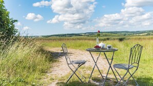 An outdoor dining setup with a table and chairs on a dirt path, surrounded by grass and a clear sky.