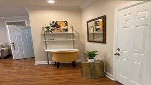 A hallway with wooden flooring, a yellow armchair, a small round table with a plant, and a white door. - Apt with utilities, wifi, a compact parking space, & on-site laundry near NYC! (Bayonne)