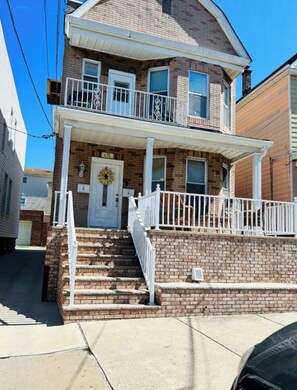 A two-story brick house with a front porch, a door with a wreath, and a number '476' on the door. - Spacious 2BR Near NYC — Parking, Patio & Transit (Bayonne)