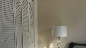A bedroom with a bed, a nightstand with a lamp, a wooden floor, and a closet with white shutters. - Apt with utilities, wifi, a compact parking space, & on-site laundry near NYC! (Bayonne)