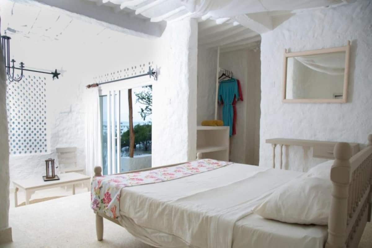 A bedroom with a bed, a window, a mirror, and a shelf with clothes.
