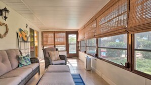 Et solbeskinnet rum med sofa, lænestol og skammel, store vinduer med persienner, og udsigt til grønne områder udenfor.