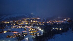 Un pueblo cubierto de nieve al caer la noche, con edificios iluminados y una vista despejada del bosque circundante.