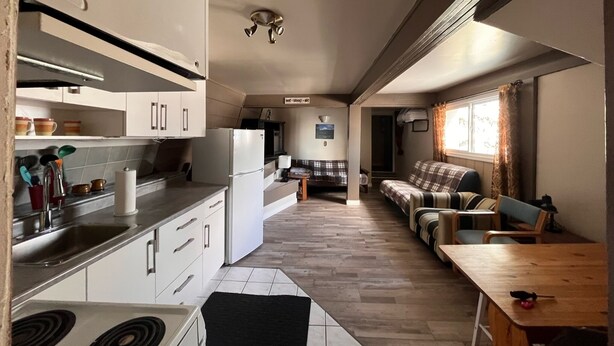 Cocina pequeña con armarios blancos, estufa y refrigerador. Cuenta con un comedor con mesa de madera y un sofá.