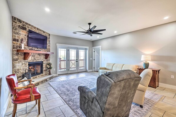 A spacious living room with a stone fireplace, a flat-screen TV, a ceiling fan, and a large window.