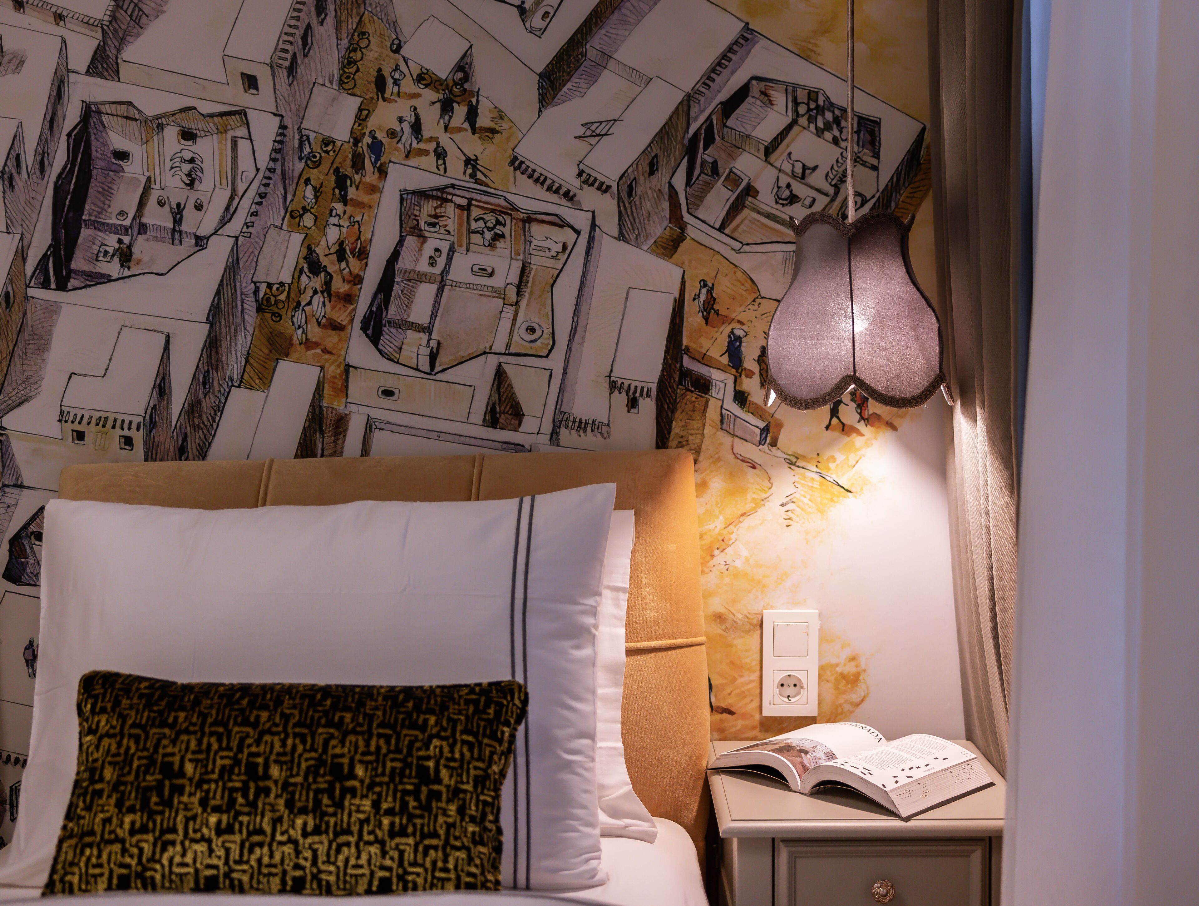 Une chambre à coucher avec un lit, des oreillers, une table de chevet sur laquelle est posé un livre ouvert, et un mur orné d’un motif décoratif.