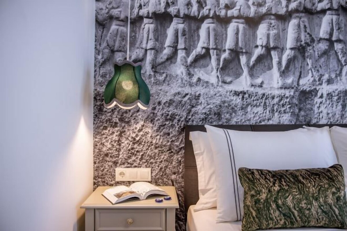Un dormitorio con cama, una mesita de noche con un libro abierto, una lámpara verde de pared y una pared con textura.