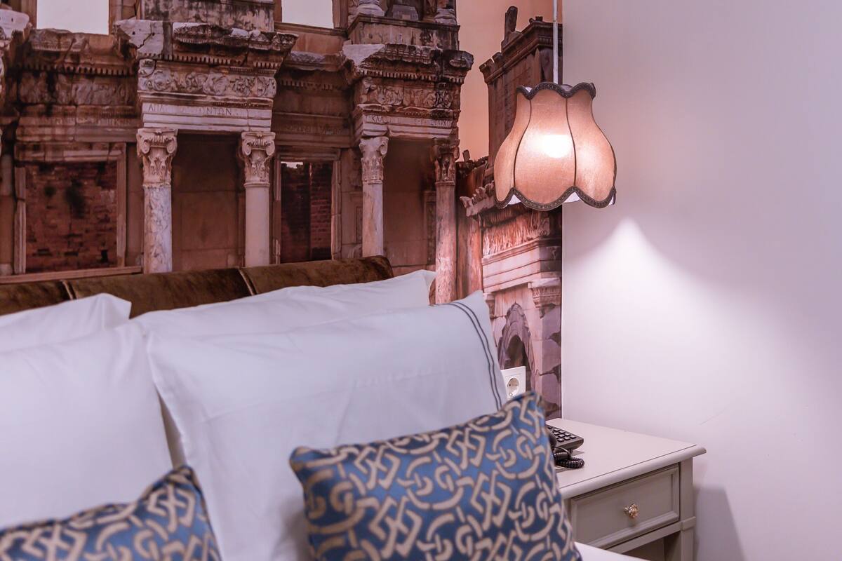 A room with a bed, pillows, a nightstand, and a wall with ancient architectural carvings.