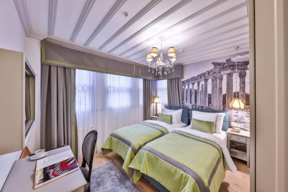 A hotel room with two beds, a desk, a chair, a chandelier, and a mural of ancient ruins.