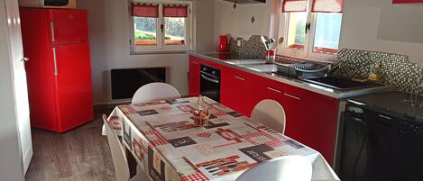 Una cocina compacta con un refrigerador rojo, una mesa de comedor con mantel a cuadros y una ventana con cortinas rojas.