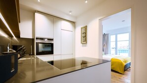 Eine moderne Küche mit einer zentralen Kochinsel, integriertem Backofen und weißen Schränken. Im Hintergrund ist ein Essbereich mit einem gelben Sofa und einem großen Fenster zu sehen.
