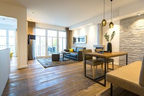 Un moderno salón con sofá, mesa de comedor y sillas. Tiene un ventanal con cortinas, una pared de piedra y vistas al exterior.