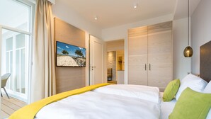 Ein modernes Schlafzimmer mit einem großen Bett, einem Fernseher an der Wand und Blick ins Freie durch eine Schiebetür.