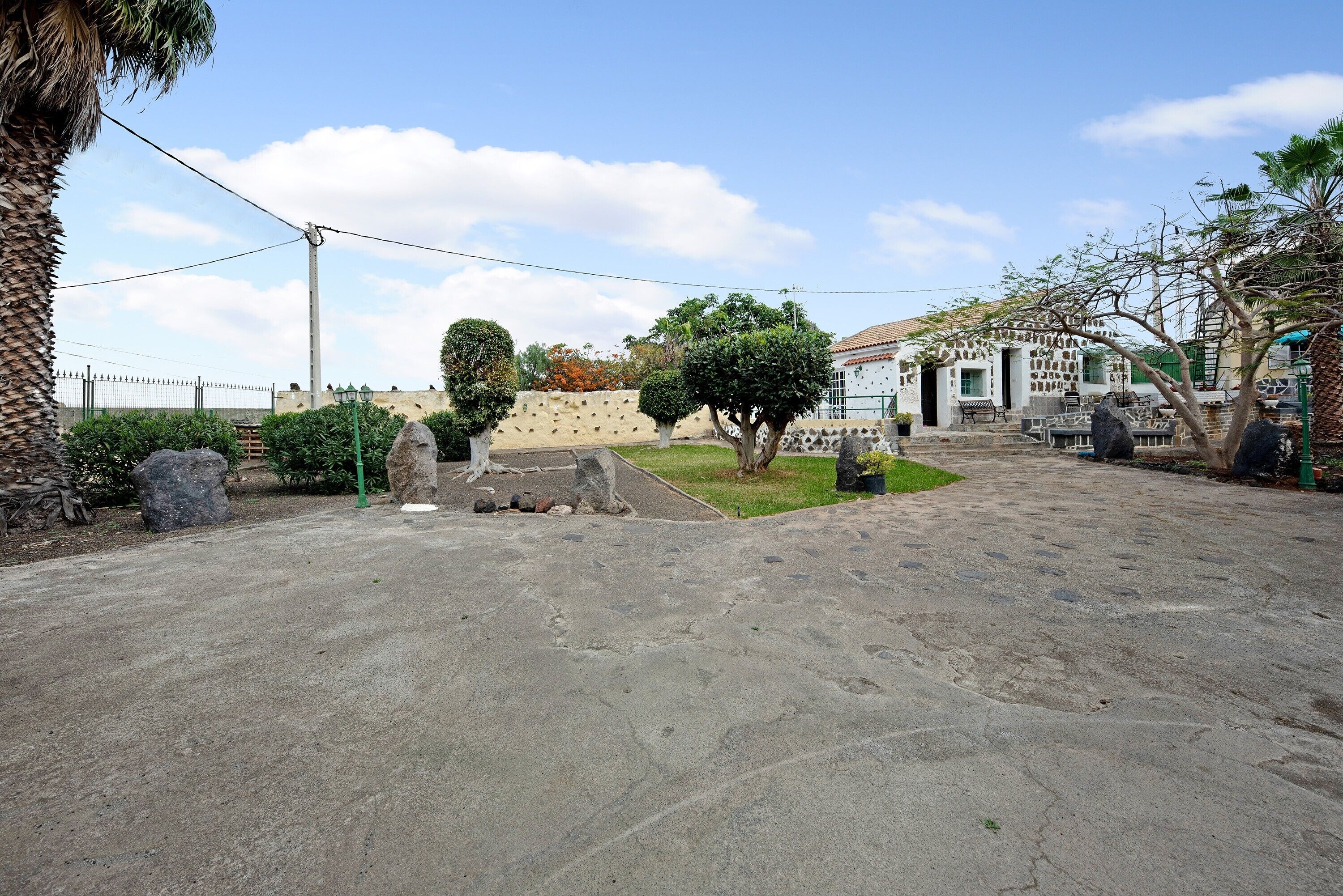 A paved courtyard with a stone wall, a palm tree, and a white building with a tiled roof.