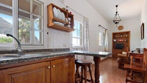 Una cocina con encimeras de granito, armarios de madera, un estante empotrado con vajilla, una zona de comedor con sillas de madera y una lámpara colgante.