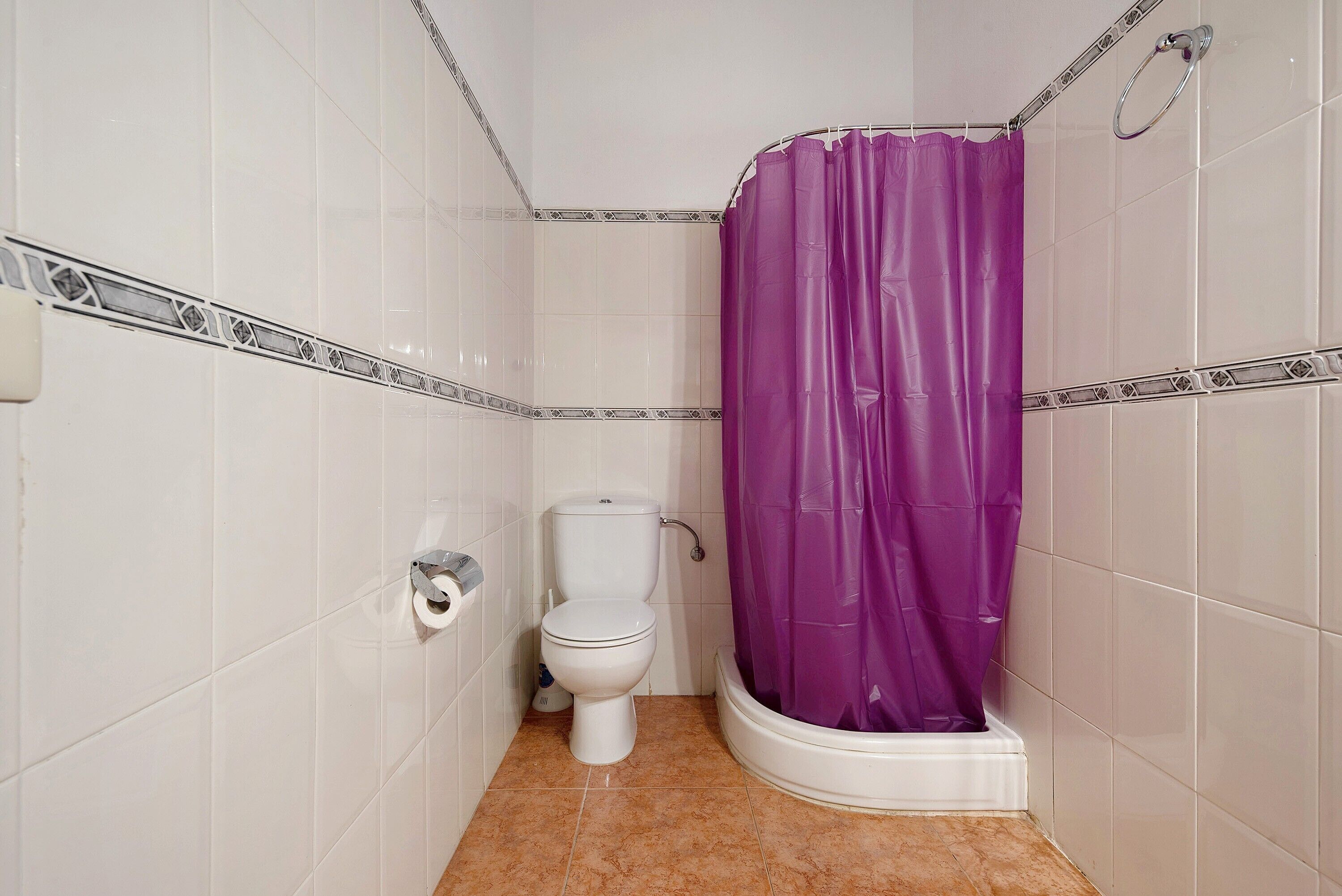 Et lille badeværelse med toilet, brusebad med lilla gardin og fliserede vægge.