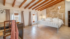 A bedroom with a white metal bed, wooden beams on the ceiling, a stone wall, and a window with curtains. - Holiday Home 'Ca Na Francesca' with Mountain View, Wi-Fi and Air Conditioning (Pollença)