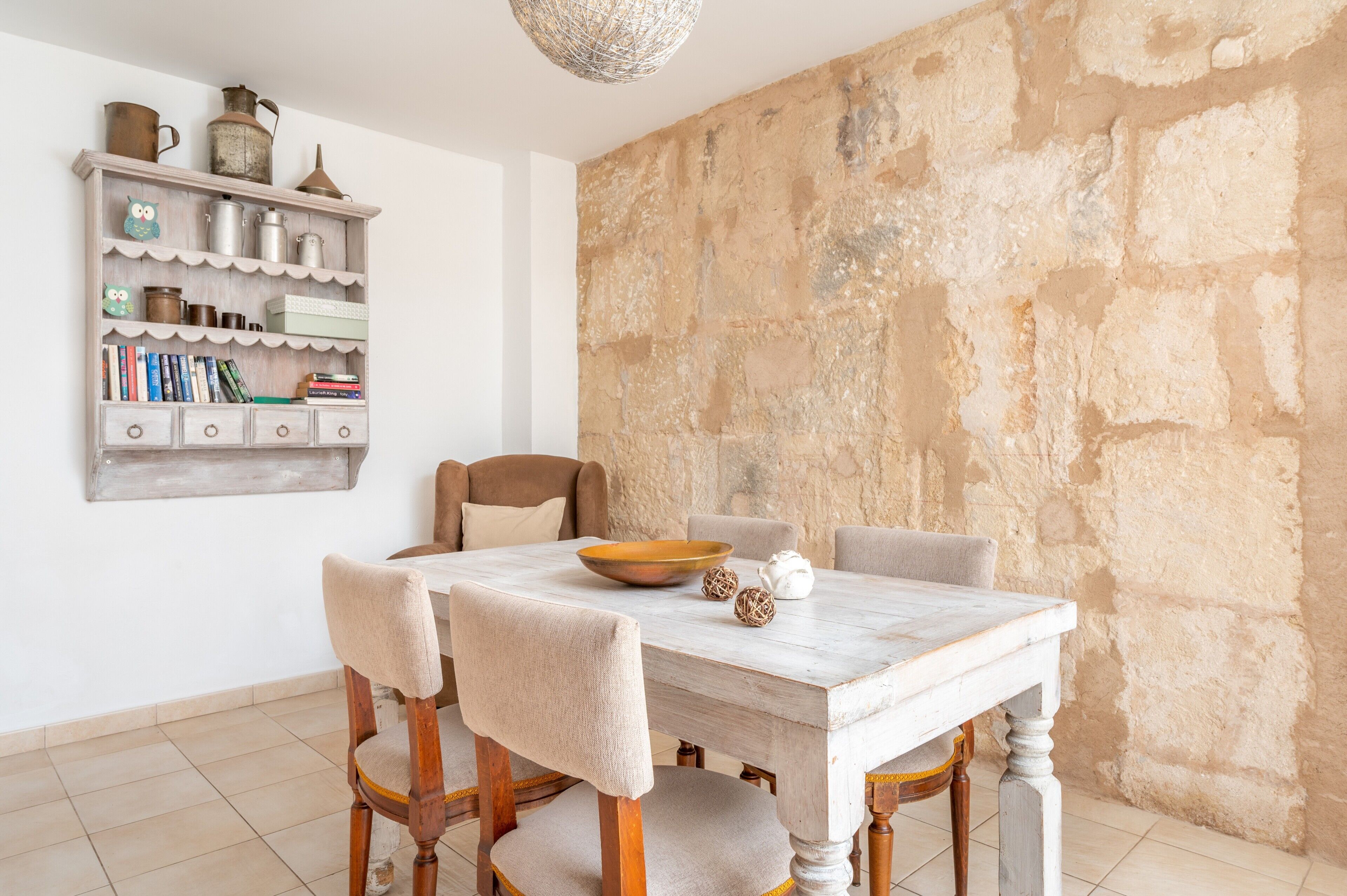 A dining area with a white table, beige chairs, a wooden bookshelf, and a stone wall.