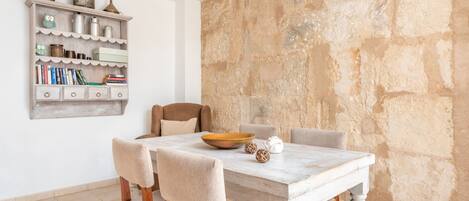 A dining area with a white table, beige chairs, a wooden bookshelf, and a stone wall.