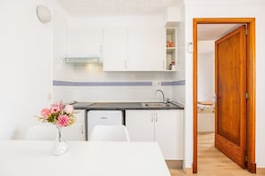 A compact kitchen with white cabinets, a built-in oven, and a sink. A dining table with chairs is positioned in front of the kitchen.