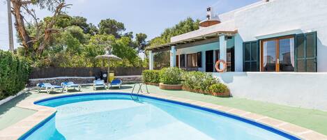 Una piscina con sillas de descanso, una pérgola y una casa con fachada blanca y contraventanas verdes.