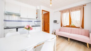 A modern kitchen with white cabinets, a dining table with chairs, a pink sofa, and a wooden door. - “Felipe 1-5” Apartment with Terrace, Shared Pool & Wi-Fi (Tourist Taxes Included) (Cala Dor)