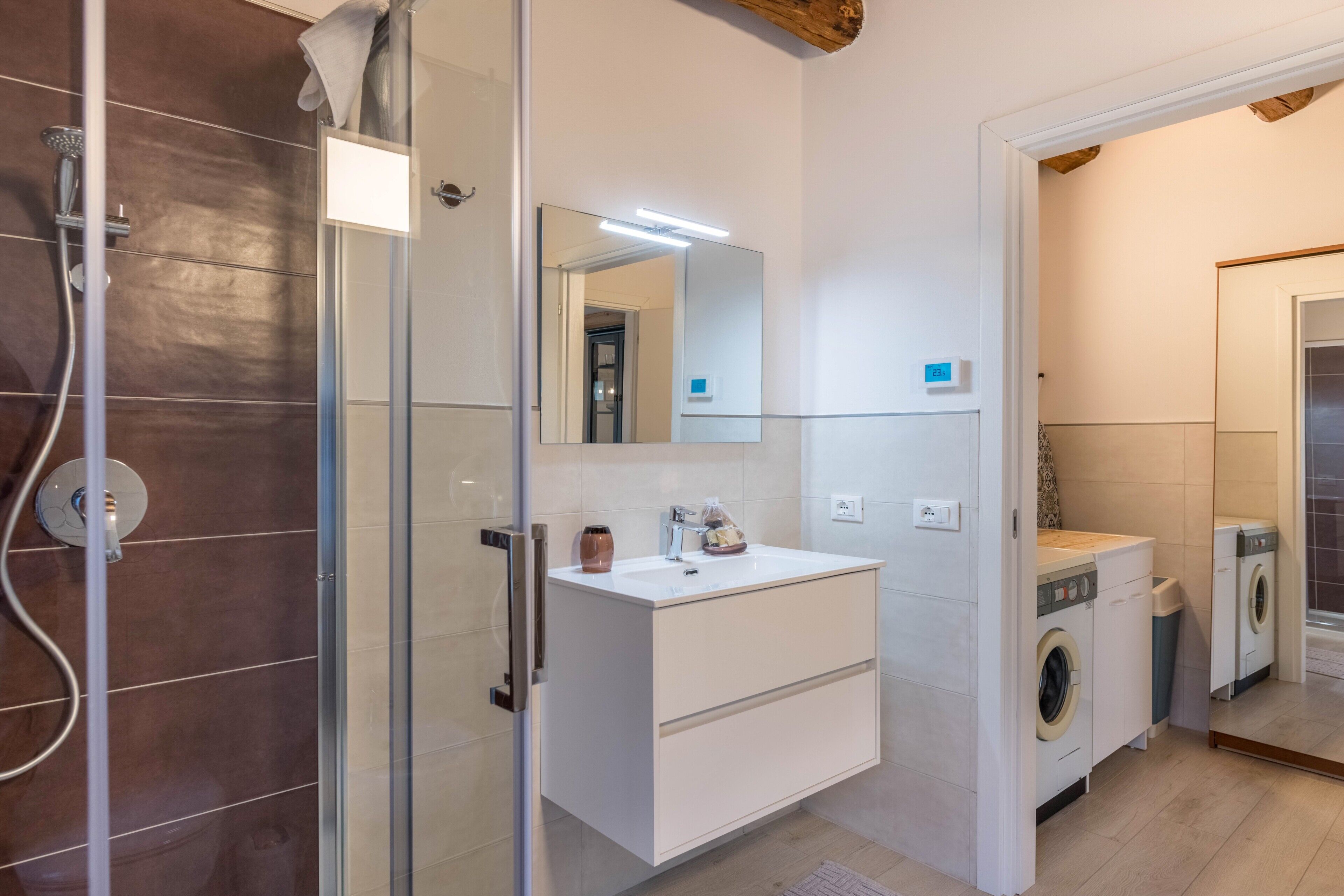 A bathroom with a glass shower enclosure, a white vanity with a mirror, a washing machine, and a toilet.