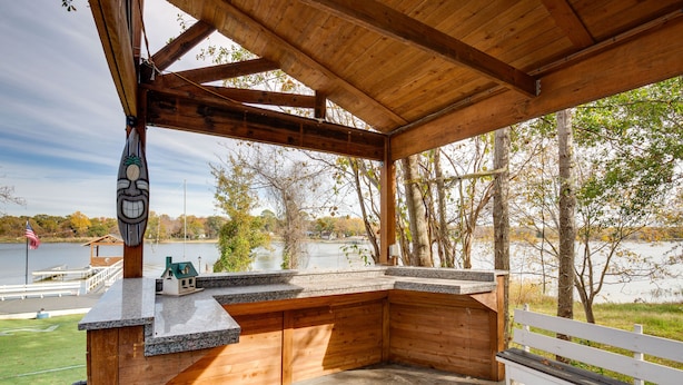 Un espacio al aire libre techado con una terraza de madera, una casita modelo y vistas a un lago y árboles.
