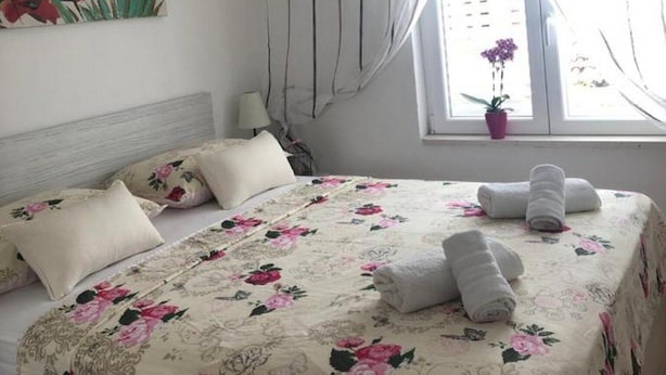 A bedroom with a floral bedspread, a single bed with pillows, a nightstand, a window with curtains, and a potted plant. - Sunone Dubrovnik (Dubrovnik)