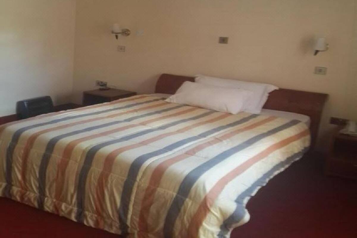 Habitación de hotel con cama, mesita de noche, lámpara y ventana con cortinas translúcidas.