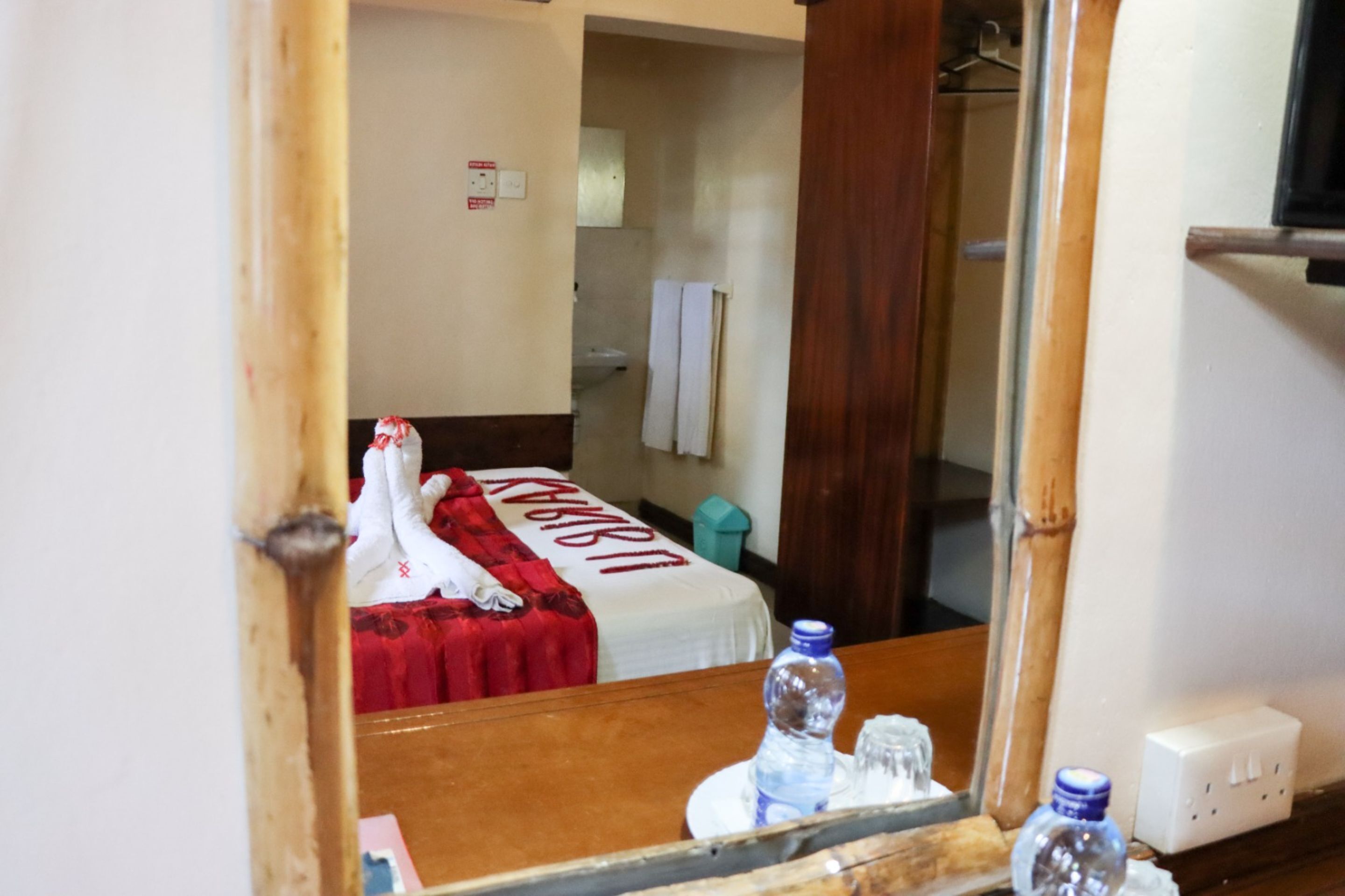A hotel room with a bed covered in a red blanket, a wooden headboard, a mirror, and a wooden table with a water bottle and glasses.