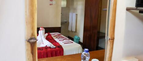 A hotel room with a bed covered in a red blanket, a wooden headboard, a mirror, and a wooden table with a water bottle and glasses.