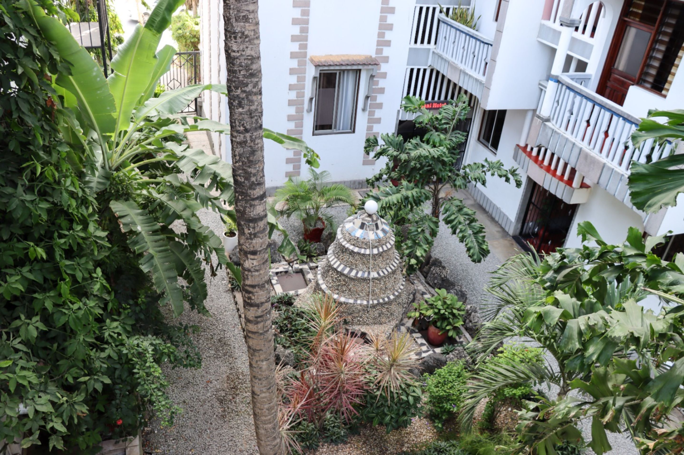 A courtyard with a decorative fountain, surrounded by various plants and a building with balconies.