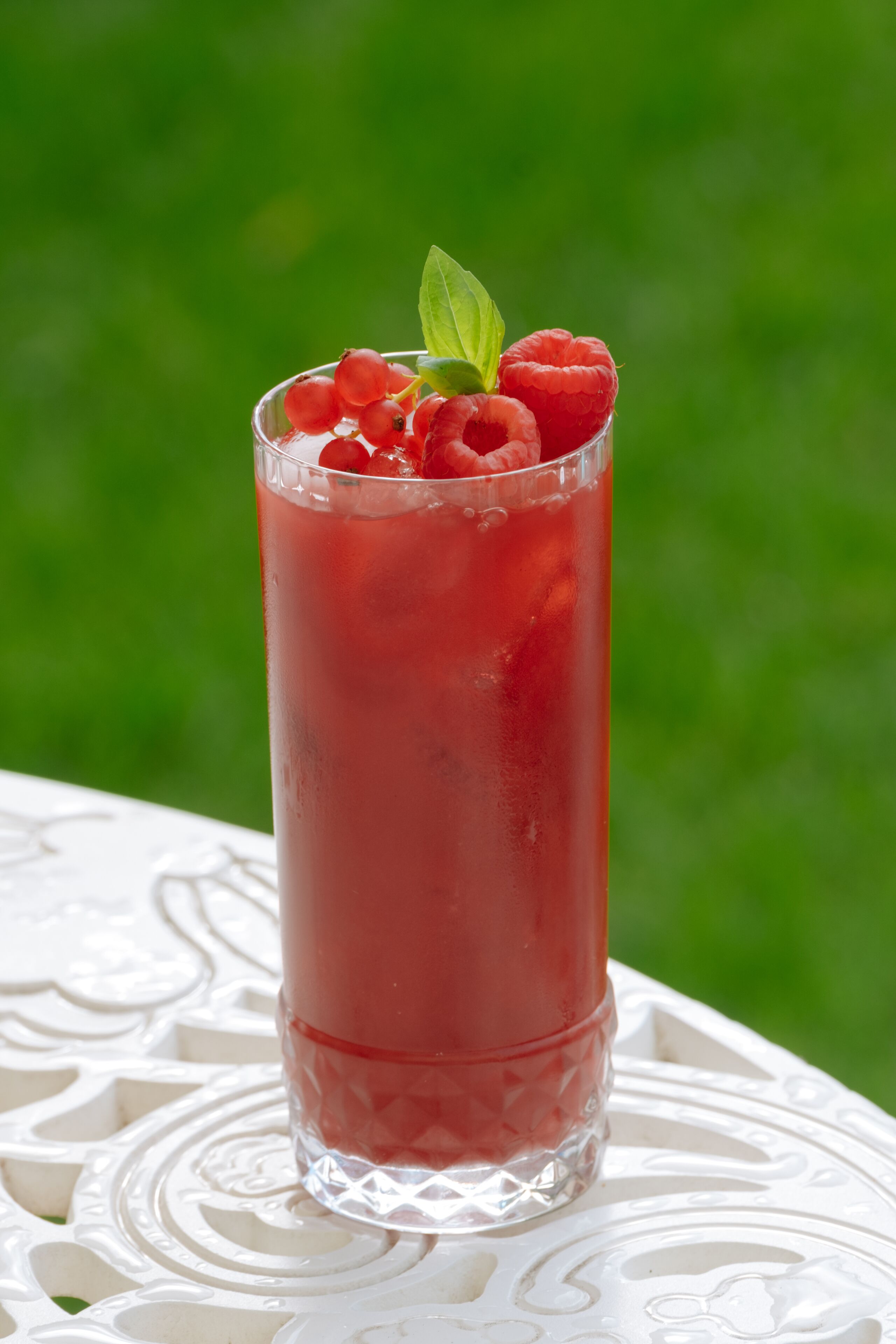 Ein hohes Glas mit einem roten Getränk, verziert mit Himbeeren und einem Minzblatt, steht auf einem weißen Tisch vor einem grünen Hintergrund.