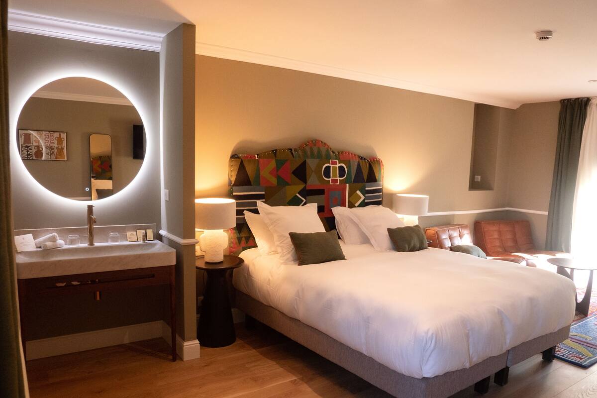 A hotel room with a large bed, a round mirror, a bedside table with a lamp, and a colorful headboard.