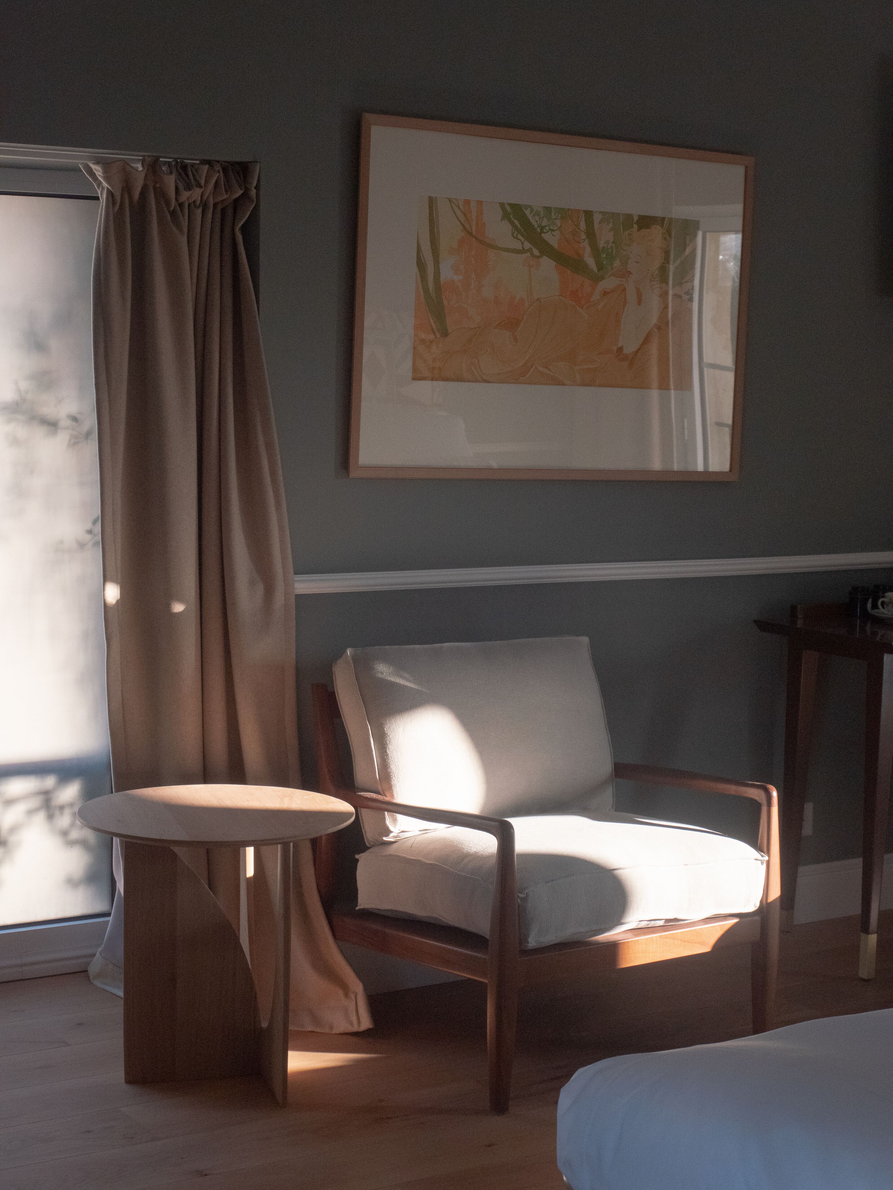 A room with a bed, a chair, a small table, a painting on the wall, and a window with curtains.