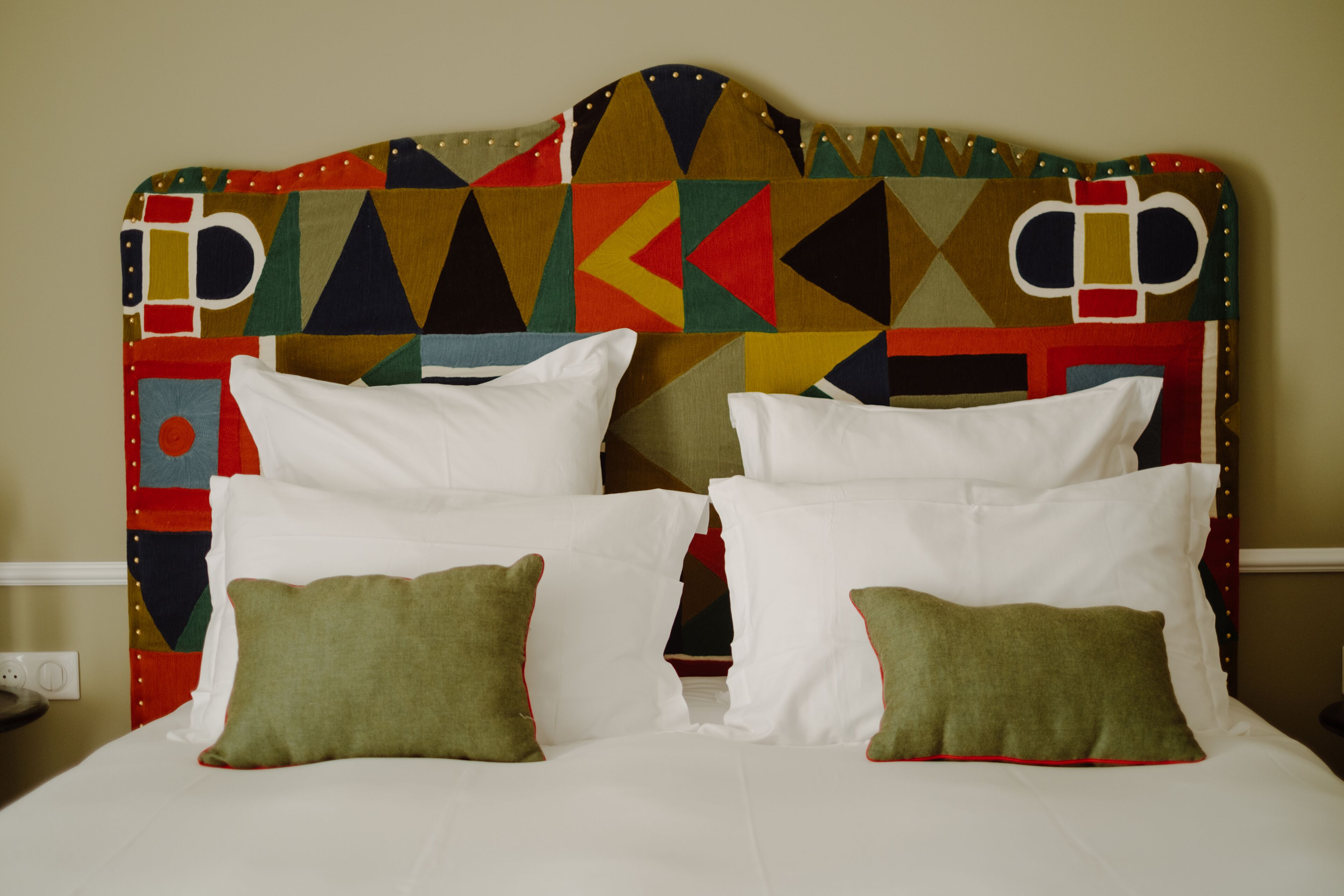 A bed with white linens and green pillows, a colorful geometric-patterned headboard, and a plain wall in the background.