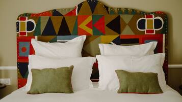 A bed with white linens and green pillows, a colorful geometric-patterned headboard, and a plain wall in the background.