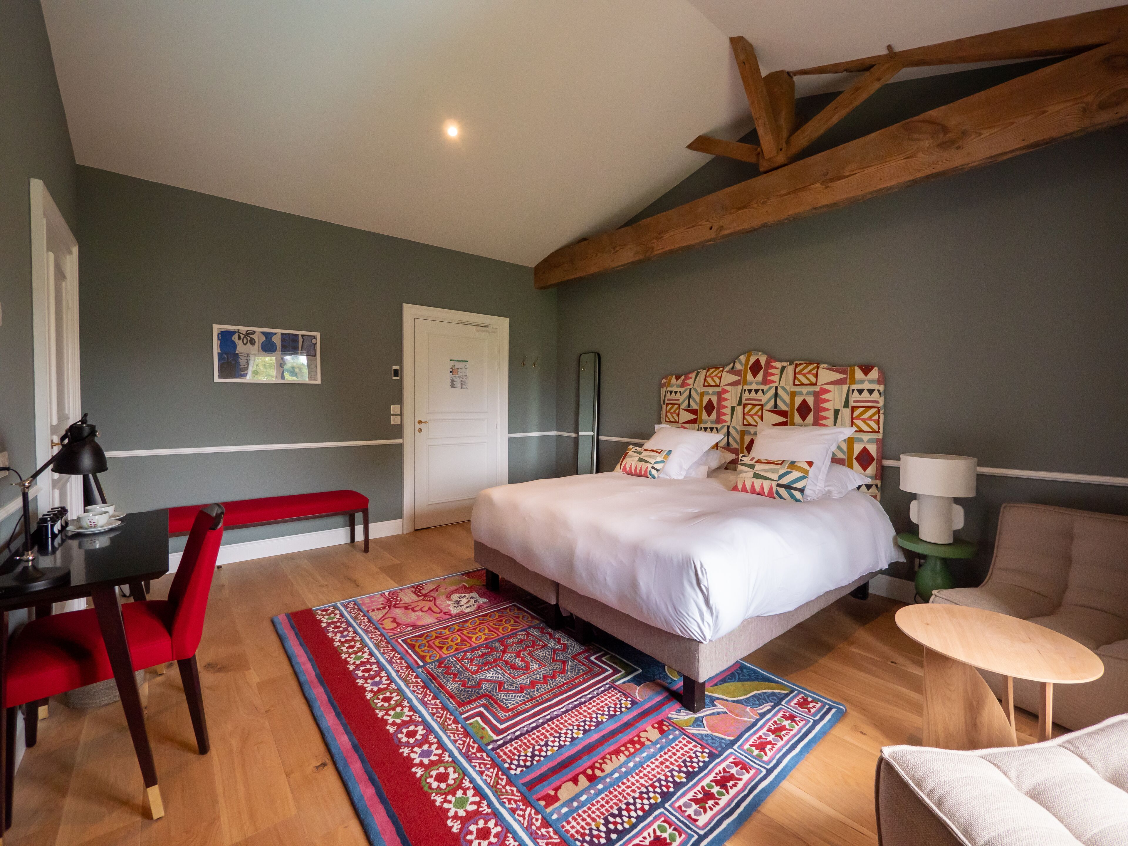 A bedroom with a bed, a desk, a chair, a small table, and a patterned rug.