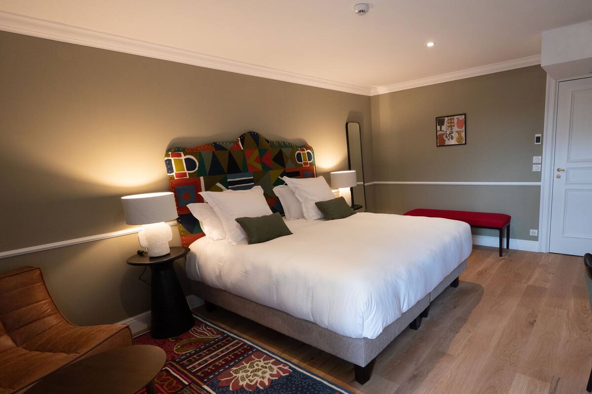 A neatly arranged bedroom with a large bed, a bedside table with lamps, a chair, and a colorful headboard.