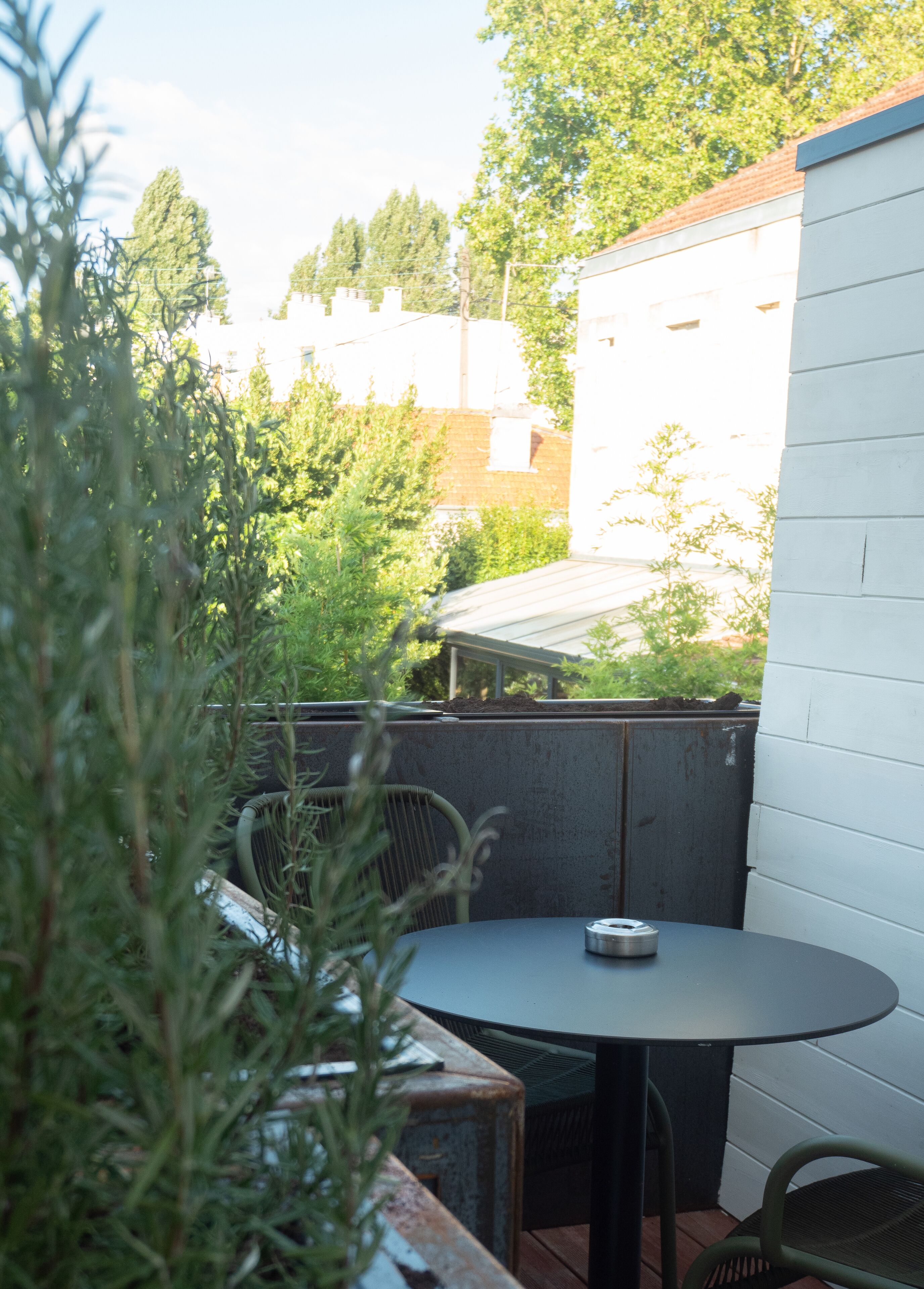 A patio area with a round table and two chairs, surrounded by greenery and a white building in the background.