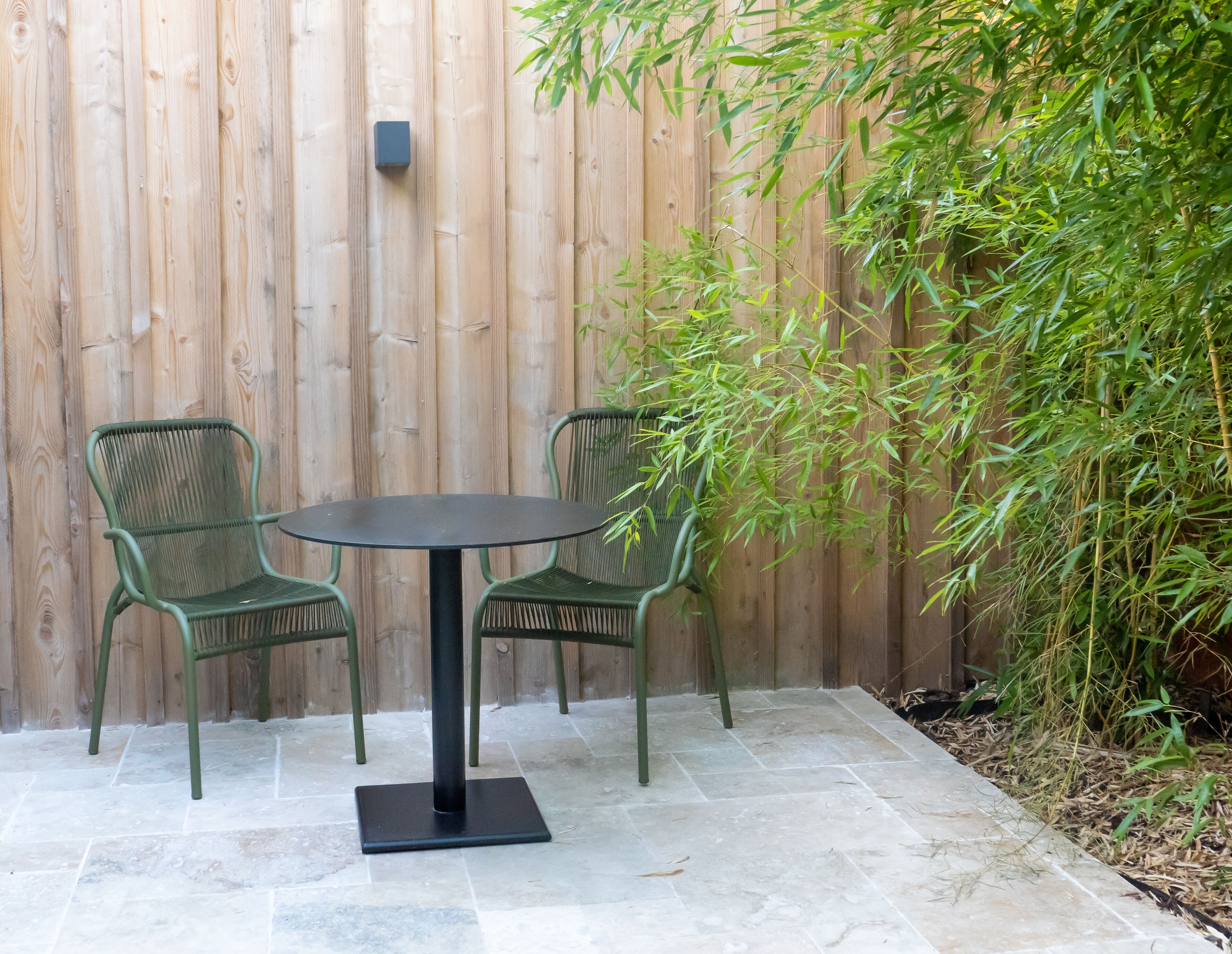 A round table with two chairs in a patio area with a wooden fence and bamboo plants.