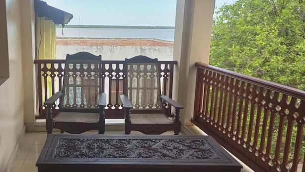 A balcony with two wooden chairs and a small table, overlooking a body of water and greenery. - New Mahrus Hotel & Rooftop Restaurant (Lamu)