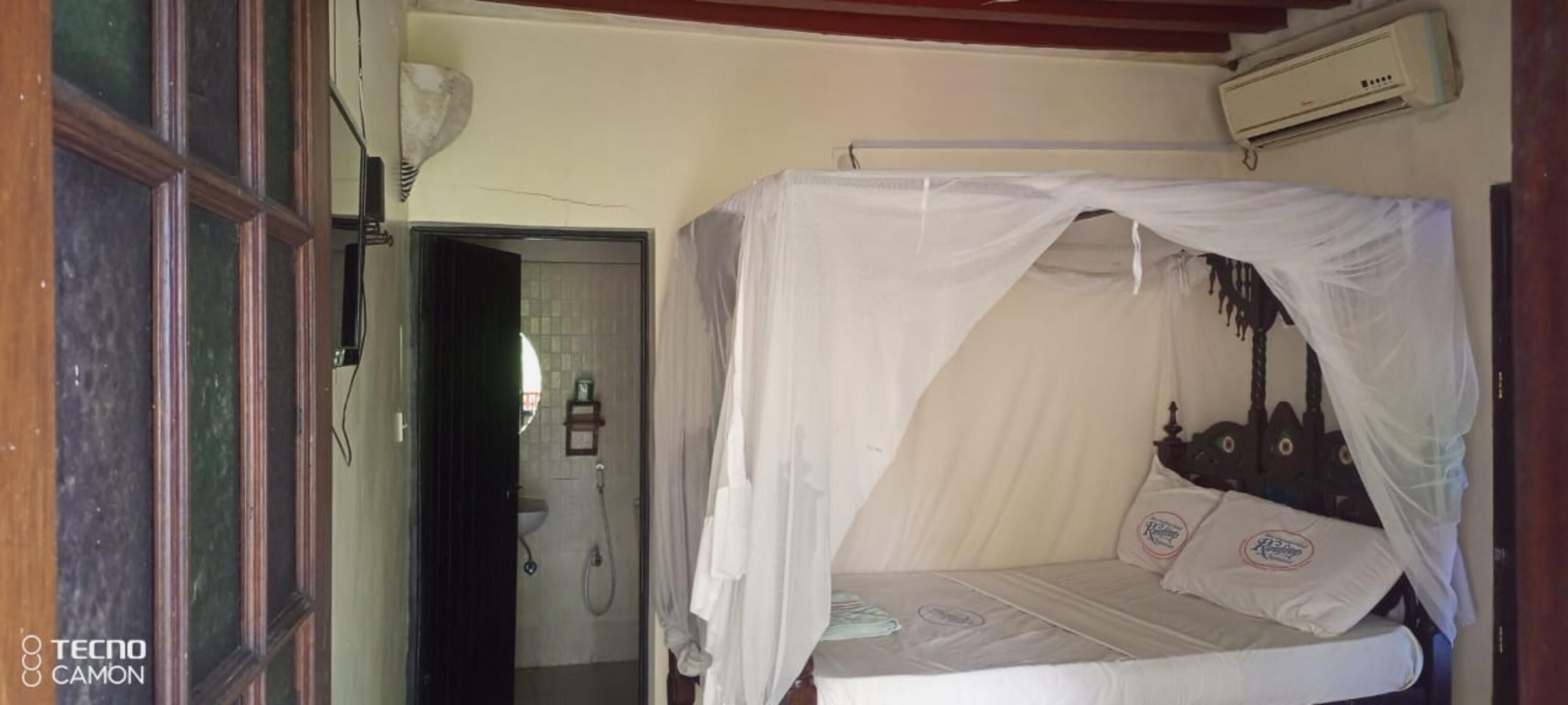 A single bed with a mosquito net, a small bedside table, and a wall-mounted air conditioner.