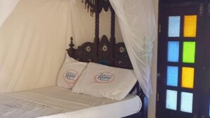 A single bed with a canopy, a wall-mounted air conditioner, and a window with colorful panes. - New Mahrus Hotel & Rooftop Restaurant (Lamu)