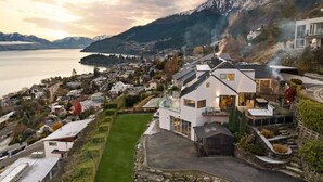 A modern house with a large driveway, a swimming pool, and a view of a lake and mountains. - Castle Cliff Lodge | Spa Pool + Sauna | Games Room (Queenstown)