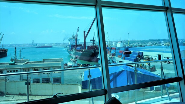 Uitzicht vanuit een raam op een gebouw, met daaronder een haven vol schepen en containers.