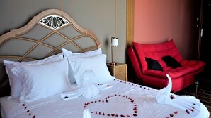 Een net opgemaakt bed met witte lakens en rozenblaadjes die in de vorm van een hart zijn gerangschikt, een rode fauteuil en een houten hoofdbord.