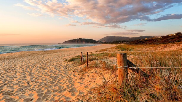 Una playa con una cerca de madera, terreno arenoso y una montaña distante.